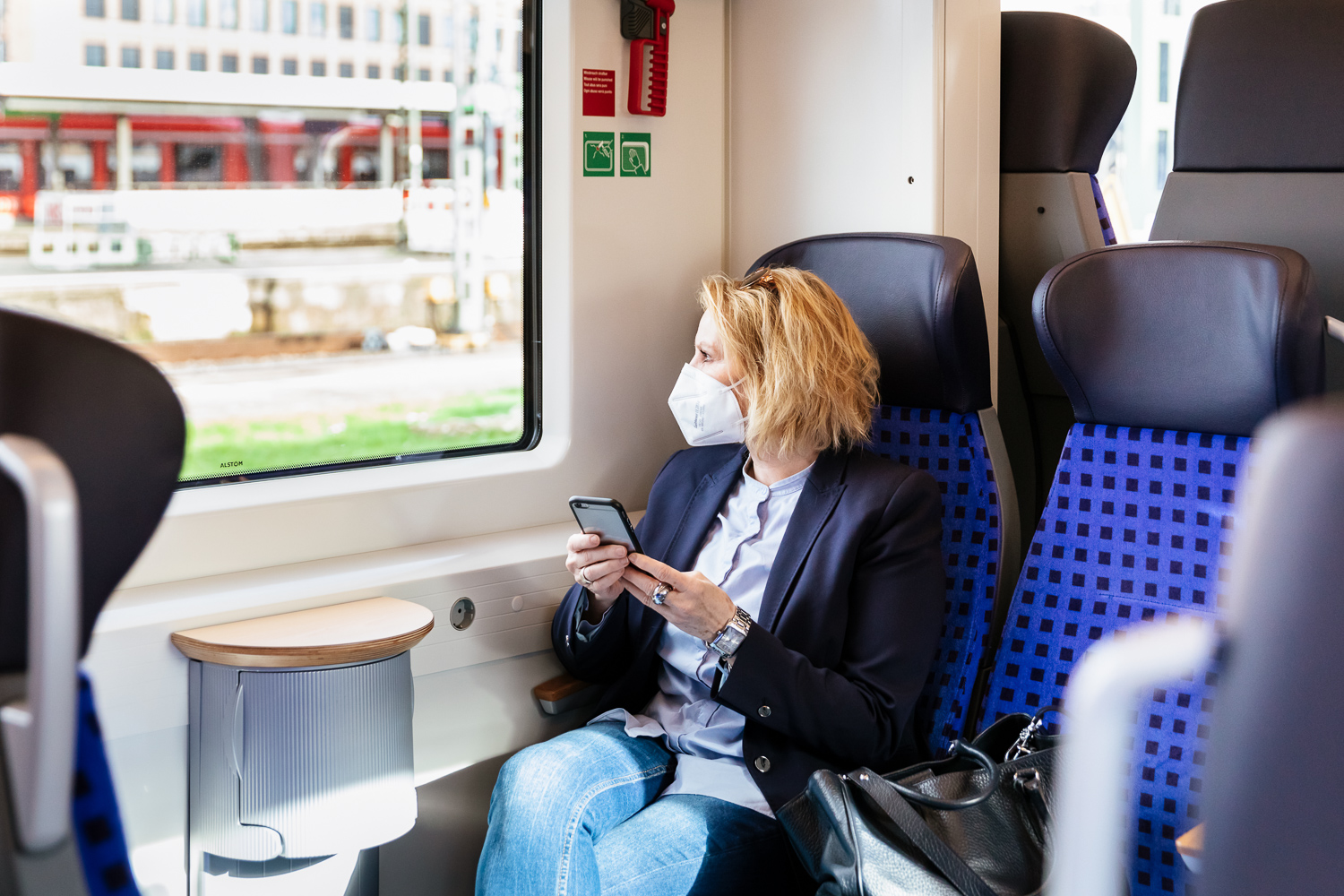 Eine Kundin mit Maske sitzt mit ihrem Smartphone in einer S-Bahn.