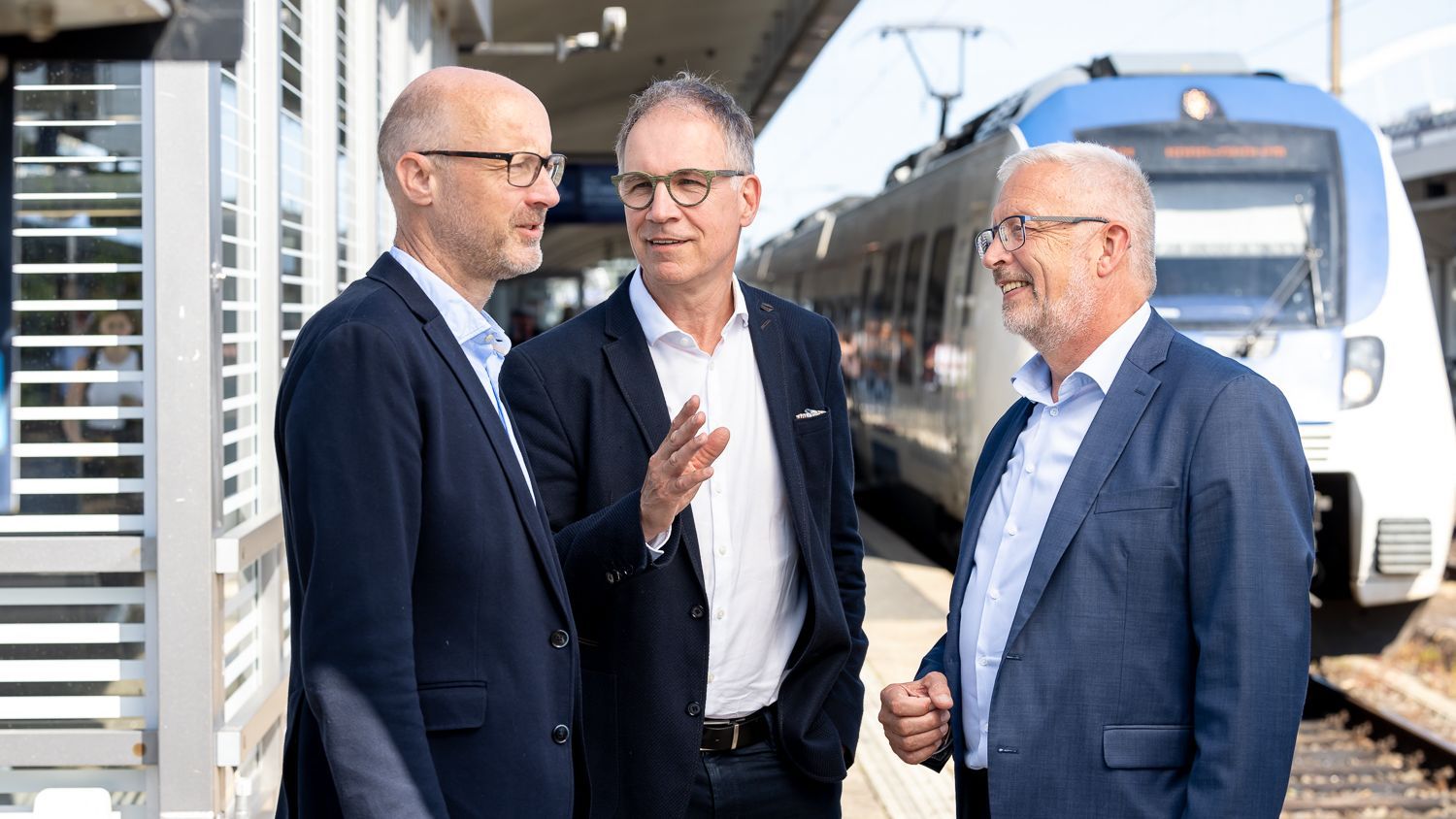 Wir sehen, wie Jens Schäfer, Norbert Reinkober und Manfred Gutfrucht zusammen auf einem Bahnsteig stehend miteinander sprechen.