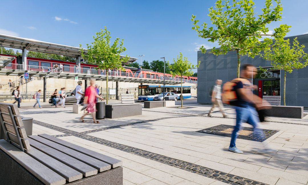 Das Bild zeigt eine sommerliche Impression der Mobilstation in Erftstadt. Mehrere Fahrgäste gehen über die weitläufige Anlage.