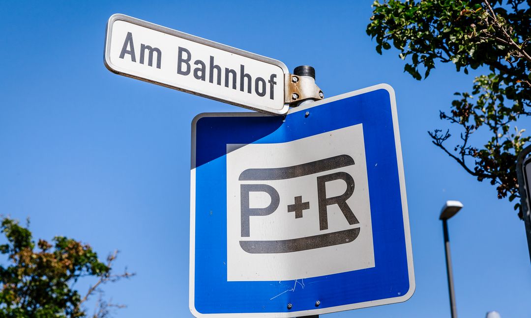 Wir sehen ein typisches Park and Ride-Schild, das direkt unterhalb eines Straßenschilds mit der Aufschrift „Am Bahnhof“ angebracht ist.